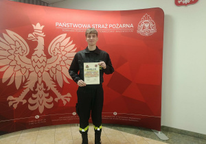 Konrad z dyplomem na tle banera Państwowej Straży Pożarnej